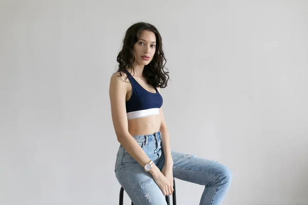 Attractive woman in sports top and jeans poses on white background.