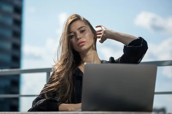 Mujer joven se sienta en la terraza con vistas a la ciudad y utiliza un ordenador portátil . — Foto de Stock