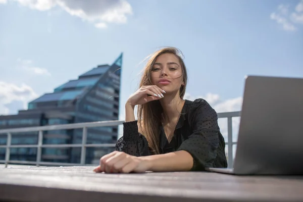Mujer joven se sienta en la terraza con vistas a la ciudad y utiliza un ordenador portátil . — Foto de Stock