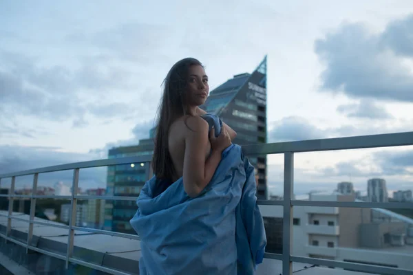 Menina nua envolto em um cobertor fica no terraço com vista para a cidade . — Fotografia de Stock