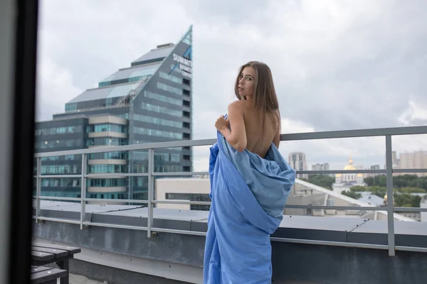 Menina nua envolto em um cobertor fica no terraço com vista para a cidade . — Fotografia de Stock