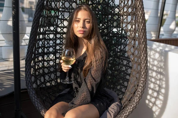 Mujer joven sentada en una silla en la terraza de un restaurante con una copa de vino . — Foto de Stock
