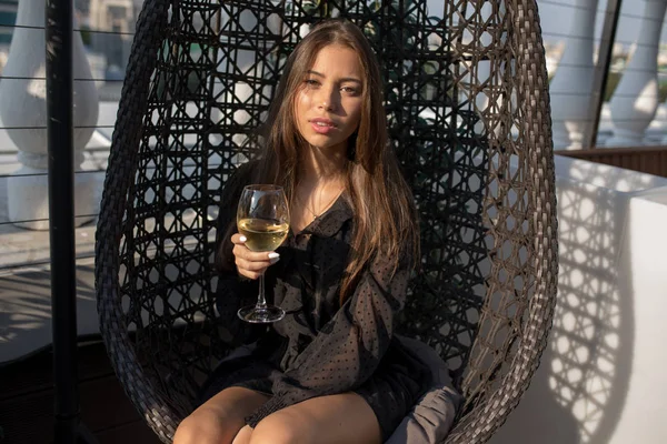 Mujer joven sentada en una silla en la terraza de un restaurante con una copa de vino . — Foto de Stock