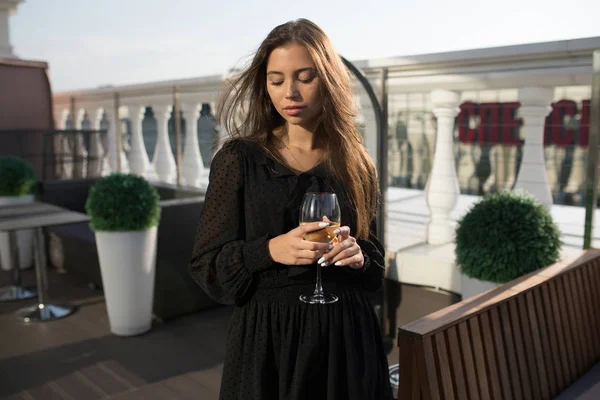 Mujer joven está de pie en la terraza de un restaurante con una copa de vino . — Foto de Stock