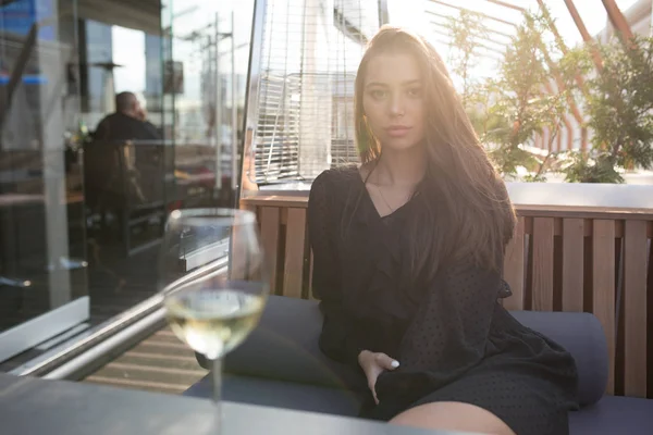 Mujer joven sentada en una mesa en la terraza de un restaurante con una copa de vino . — Foto de Stock