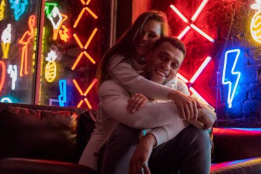 Neon lambaları arka plan üzerinde kucaklayan mutlu çok ırklı çift oturur.