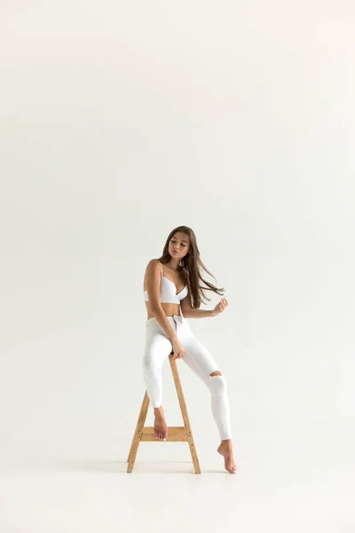 Jovem atraente em sutiã branco e jeans senta-se no fundo branco . — Fotografia de Stock