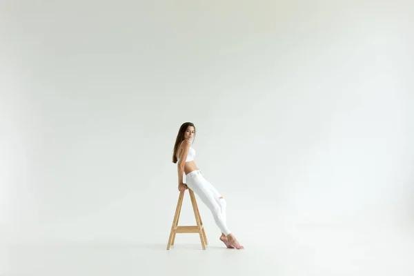 Привлекательная молодая женщина в белом лифчике и джинсах сидит на белом фоне . — стоковое фото