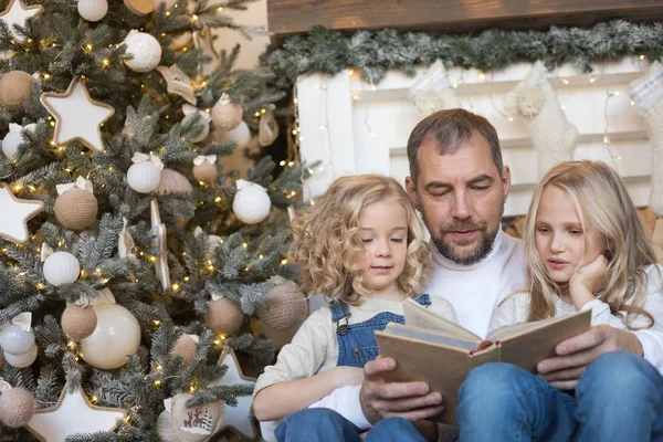 Papá lee a las hijas un libro cerca del árbol de Navidad . — Foto de Stock