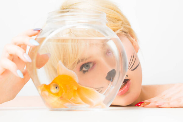 Portrait of a girl with cat make-up looks at an aquarium with a goldfish.