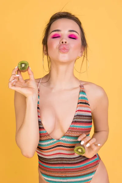 Emotional girl in a swimsuit holding a kiwi on a yellow background. — Stock Photo, Image