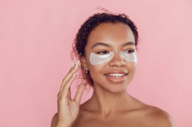 Pembe arka planda gözlerinin altında kolajen pedleri olan Afro-Amerikalı genç bir kadın..