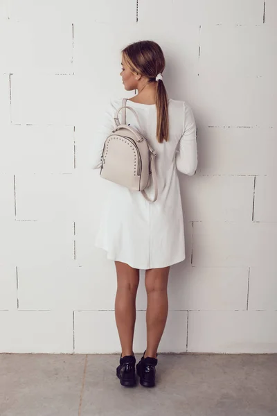 View from the back to a girl in a light dress and a backpack on her shoulder.