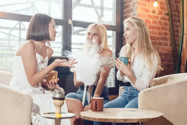 Három lány barátnő ül egy kávézóban, beszélgetnek és szívnak egy vízipipát.. — Stock Fotó
