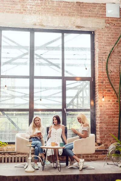 Drei Freundinnen sitzen in einem Café, unterhalten sich und rauchen eine Wasserpfeife. — Stockfoto