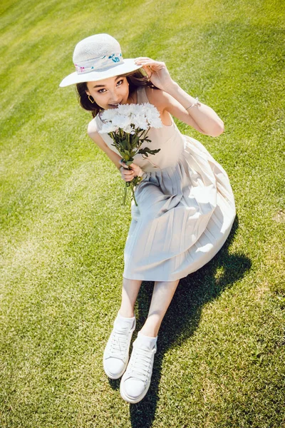 Atractiva joven en un vestido ligero y sombrero se sienta en la hierba con un ramo de flores en sus manos . —  Fotos de Stock