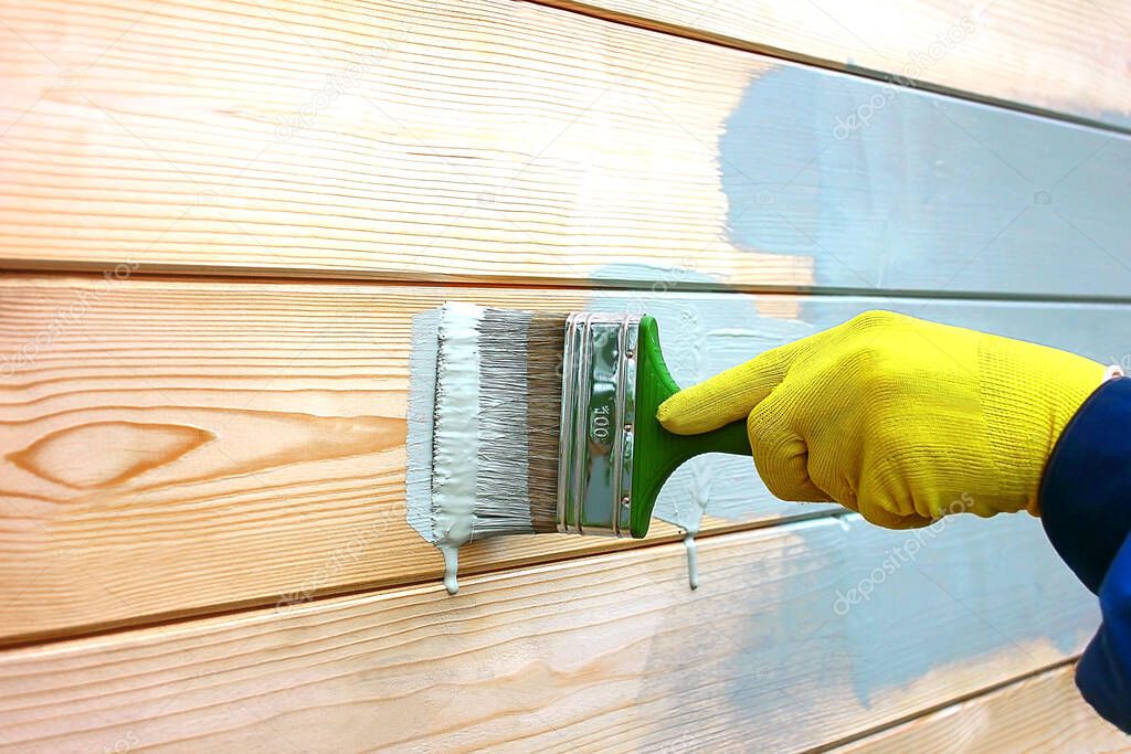 Male work hand in a yellow work glove paints the boards with a green brush on the wooden wall of the house, with acrylic blue-gray paint.