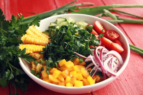 White plate with cut vegetables for a vegan salad on the red wooden table. Colorful photo. Corn, cherry tomatoes, bell pepper, onion, cucumber and garden herbs.