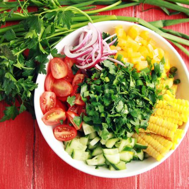 Beyaz tabak kırmızı ahşap masa üzerinde bir vegan salata için kesilmiş sebzeler ile. Renkli fotoğraf. Mısır, domates, dolmalık biber, soğan, salatalık ve Bahçe otlar. Yakın çekim. Tonda fotoğraf. Üstten görünüm.