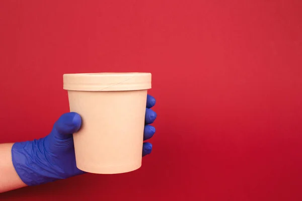 Courier hand in blue disposable protective gloves on burgundy red background, holding round paper food container - cardboard cup for soup, ice cream or other dishes to-go. Copy space for text. Mockup