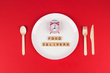 Beyaz plaka, pembe alarm saati, ahşap çatal bıçak seti ve tahta tuğlalar üzerindeki gıdalar, kırmızı bordo renkli arka plan ve kopyalama alanı. Yaratıcı gıda dağıtım konsepti, seçici odaklanma