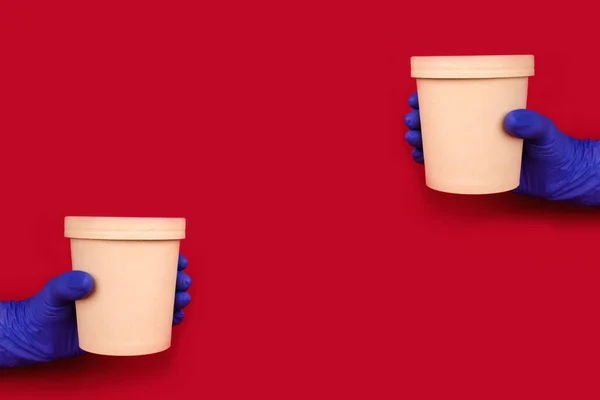 Composition with hands wearing blue disposable protective gloves on red background holding round paper food container - cardboard cup for soup or other dishes takeaway. Copyspace, mockup