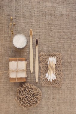 Doğal banyo aletleriyle dikey kompozisyon, sürdürülebilir yaşam tarzı konsepti. Ahşap diş fırçaları, bambu çubukları, organik sabun, diş pudrası, kumaş zemin üzerinde jüt bezi ve fotokopi alanı