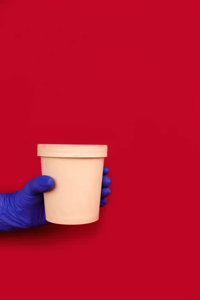 Composición vertical con la mano con guantes protectores desechables azules, sobre fondo rojo sosteniendo contenedor de papel redondo de comida taza de cartón para sopa u otros platos para llevar. Copyspace, maqueta — Foto de Stock