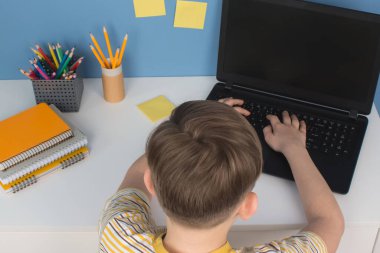 Boy using laptop computer for online studying during quarantine, sitting at the table agains blue wall. Homeschooling, online training classes or education technology concept clipart