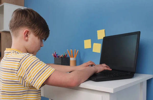 Boy using black desktop computer for online studying during quarantine, sitting at the table agains blue wall. Homeschooling, online training classes or education technology concept