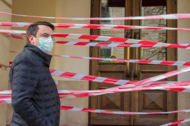 Coronavirus ya da Covid-19 pandemisi sırasında tek kullanımlık koruyucu maske takan yakışıklı Avrupalı genç adam, kapalı restoran ya da dükkan kapısının yanında kırmızı bantlı..
