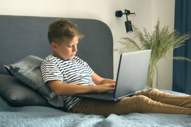 Çocuk ev ödevi yapmak için dizüstü bilgisayarını kullanıyor, modern iç mekanlarda yatağında oturuyor. Karantina ya da kişisel eğitim konsepti sırasında internetten öğrenim görmek. Kişisel eğitim planı