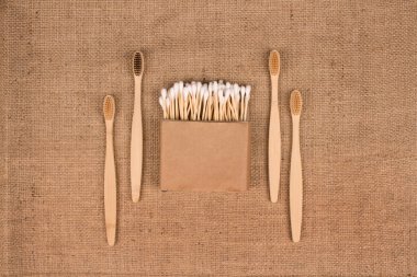 Kraft kağıt kutusunda pamuk tomurcukları ve doğal çuval bezi arkaplanında doğal bambu diş fırçaları - sıfır atık banyo esaslı. Sürdürülebilir yaşam tarzı konsepti. Model ve Kopyalama Alanı