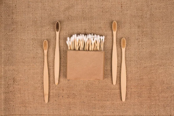 Kraft kağıt kutusunda pamuk tomurcukları ve doğal çuval bezi arkaplanında doğal bambu diş fırçaları - sıfır atık banyo esaslı. Sürdürülebilir yaşam tarzı konsepti. Model ve Kopyalama Alanı
