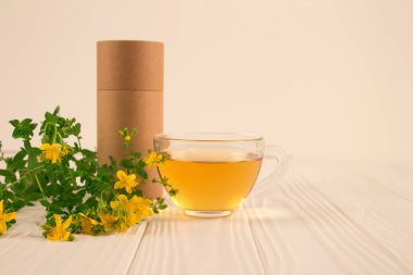 Şeffaf fincanda taze bitkisel Hypericum çayı, sarı çiçekler ve beyaz ahşap masada kağıt ambalaj tüpü - alternatif tıp konsepti. Tıpta faydalı bir bitki Hypericum. Kopyalama ve modelleme