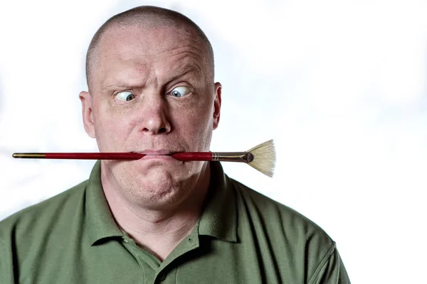 Brutal hombre calvo sostiene un cepillo de arte en su boca y hace una cara — Foto de Stock