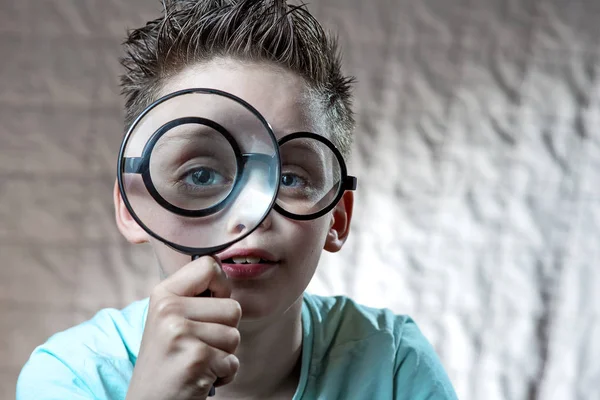 Αγόρι σε ένα ελαφρύ μπλουζάκι και γυαλιά κοιτάζοντας σε ένα μεγάλο μεγεθυντικό φακό — Φωτογραφία Αρχείου