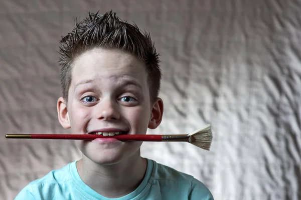 Niño en una camiseta ligera sosteniendo un cepillo de arte en su boca y sonriendo — Foto de Stock