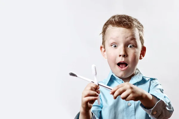 Dreng i en blå skjorte holder tandbørster og glæder sig på en lys baggrund - Stock-foto