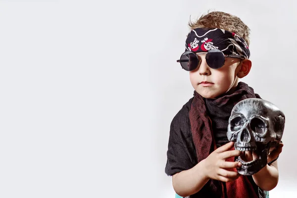 Un niño rockero en gafas negras, bufanda, bandana y con un cráneo en las manos sobre un fondo claro — Foto de Stock