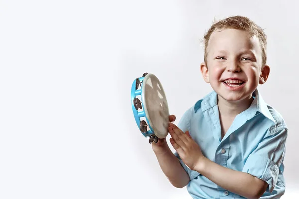 Un niño alegre con una camisa azul sosteniendo una pandereta y sonriendo sobre un fondo claro — Foto de Stock