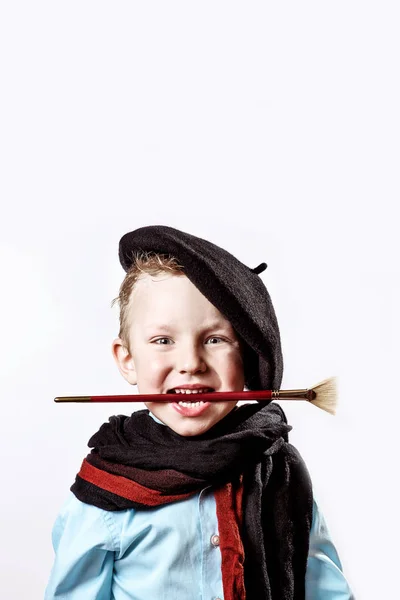 Dreng kunstner i sort baret, tørklæde og med en børste i munden på en lys baggrund - Stock-foto