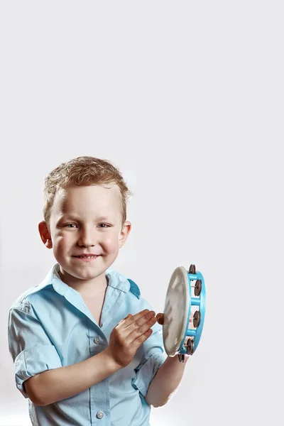 En munter dreng i en blå skjorte, der holder en tamburin og smiler på en lys baggrund - Stock-foto