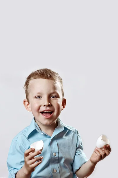 Dreng i en lys skjorte holder en skal fra ægget og smiler på en lys baggrund - Stock-foto