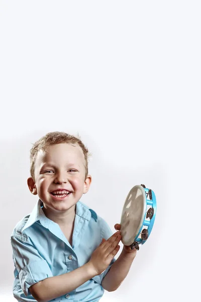 En munter dreng i en blå skjorte, der holder en tamburin og smiler på en lys baggrund - Stock-foto