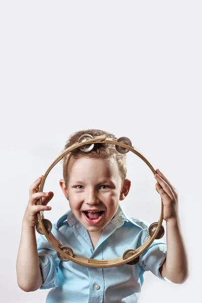 Un ragazzo allegro con una camicia blu che regge un tamburello e sorride su uno sfondo leggero — Foto Stock