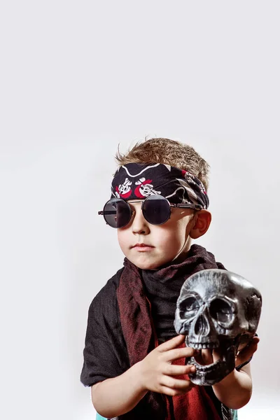 Un niño rockero en gafas negras, bufanda, bandana y con un cráneo en las manos sobre un fondo claro — Foto de Stock