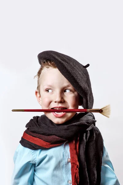 Ragazzo artista in berretto nero, sciarpa e con un pennello in bocca su uno sfondo chiaro — Foto Stock