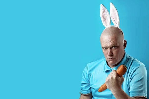 Hombre calvo y brutal con camiseta ligera y orejas de conejo. En sus manos sostiene la zanahoria como una mancuerna . — Foto de Stock