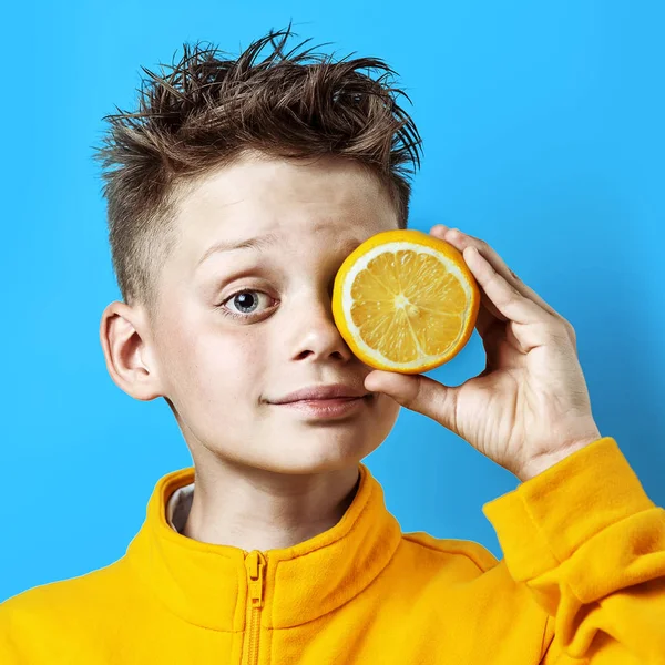 Dreng i en lys gul jakke med en citron i hånden på en blå - Stock-foto